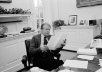 black and white photo of former president jimmy carter in a sweater behind a desk looking at papers.